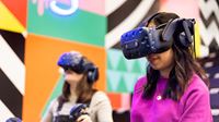 Two women wearing virtual reality headsets with colourful geometric patterns on the wall in the background.