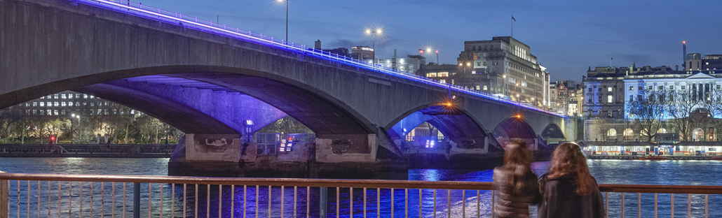 Two people look at Waterloo Bridge illuminated as part of Illuminated River