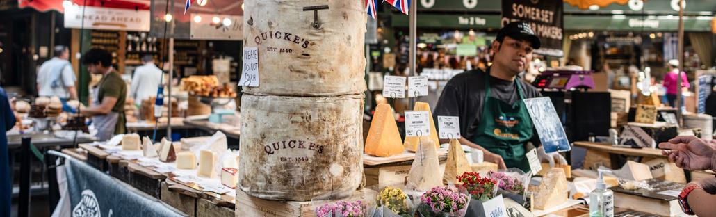 Borough Market food stalls with cheese, flowers, bread and more, and traders ready to welcome customers.