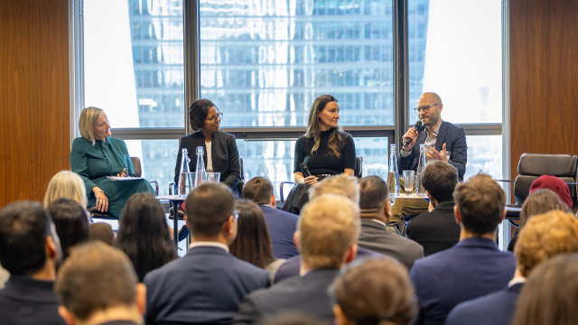 A conference room full of people listening to four speakers in a panel with a skyscraper visible from a window in the background.