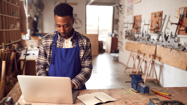 A man wearing blue overalls and a flannel shirt working on a laptop with a workshop in the background.