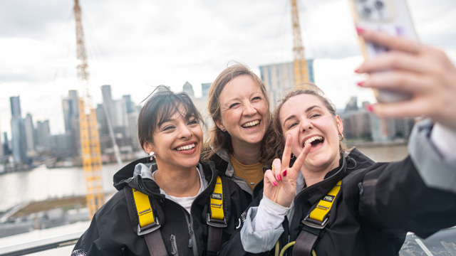Three women smiling while taking a selfie with a panoramic view of London in the background.