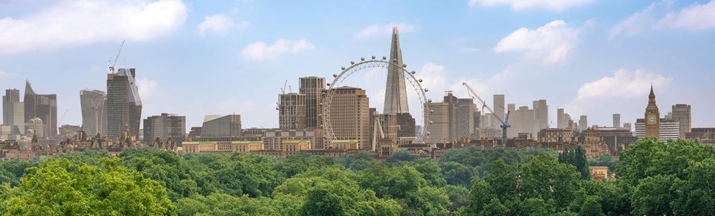 A daytime view of the London skyline in the background, with the Shard, Eye of London and Big Ben visible, and green trees in Hyde Park in the foreground.