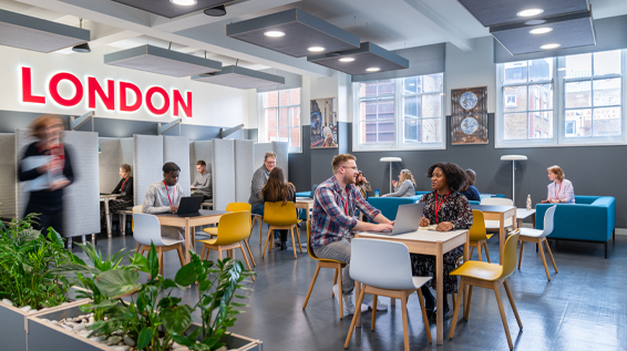 A view of an office collaboration area with groups of people sitting at tables and people working in booths.