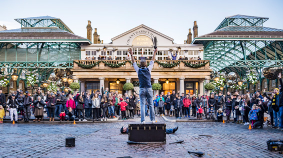 A day time view of a street performer standing on top of a stage performing to a crowd of people. 