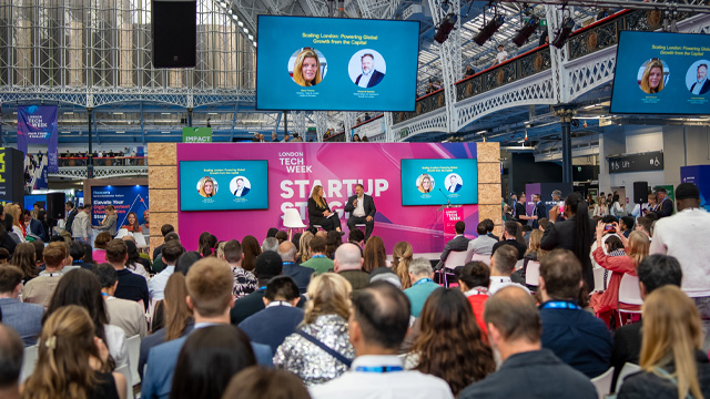 A stage at a business conference with two panelists speaking to a crow of people.