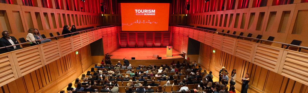 A view of a theatre looking over rows of seats filled with an audience looking towards the stage with a red screen that reads Tourism Means Business.