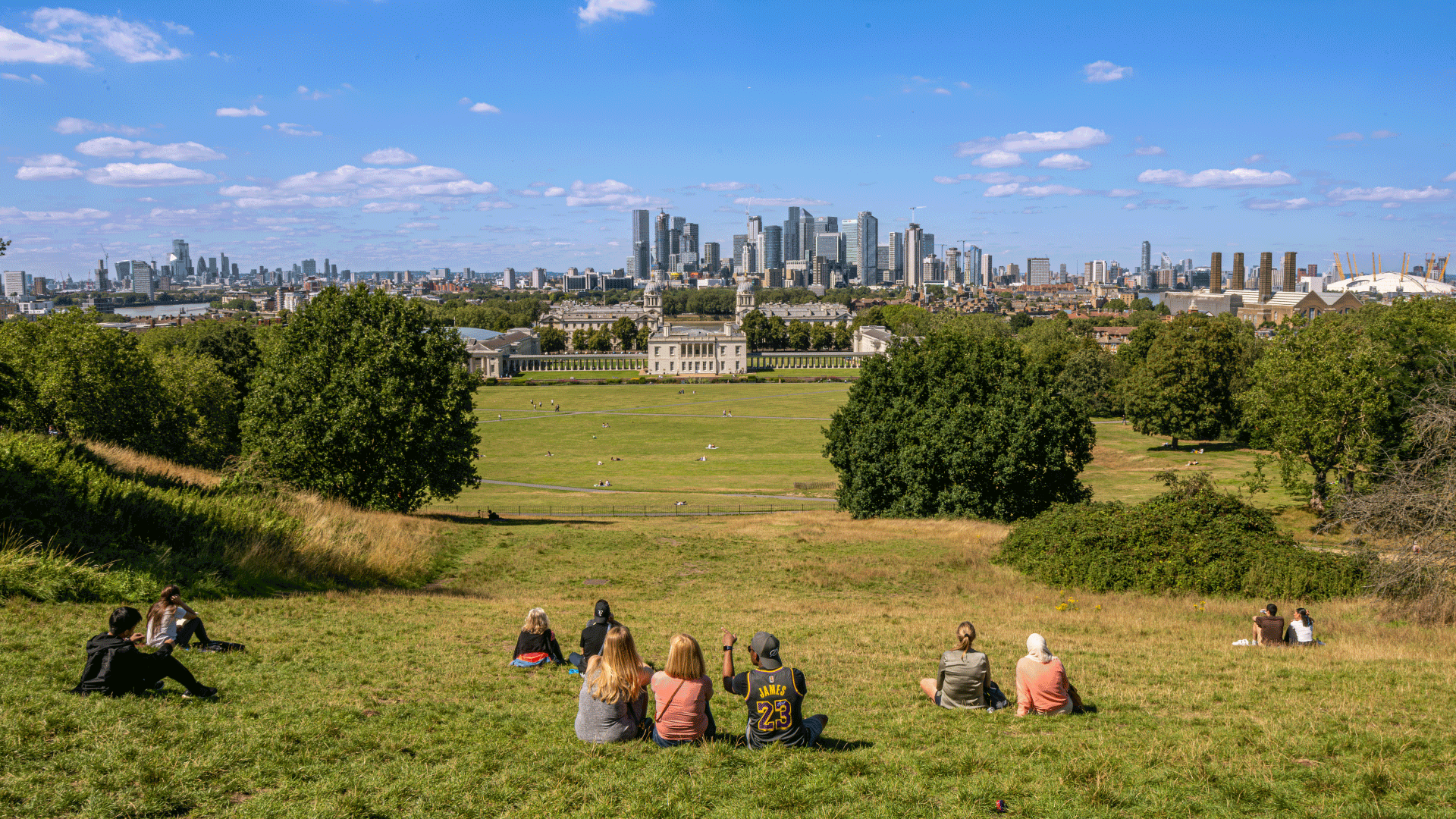 Groups of people sat on the grass looking at the view of London on a sunny day